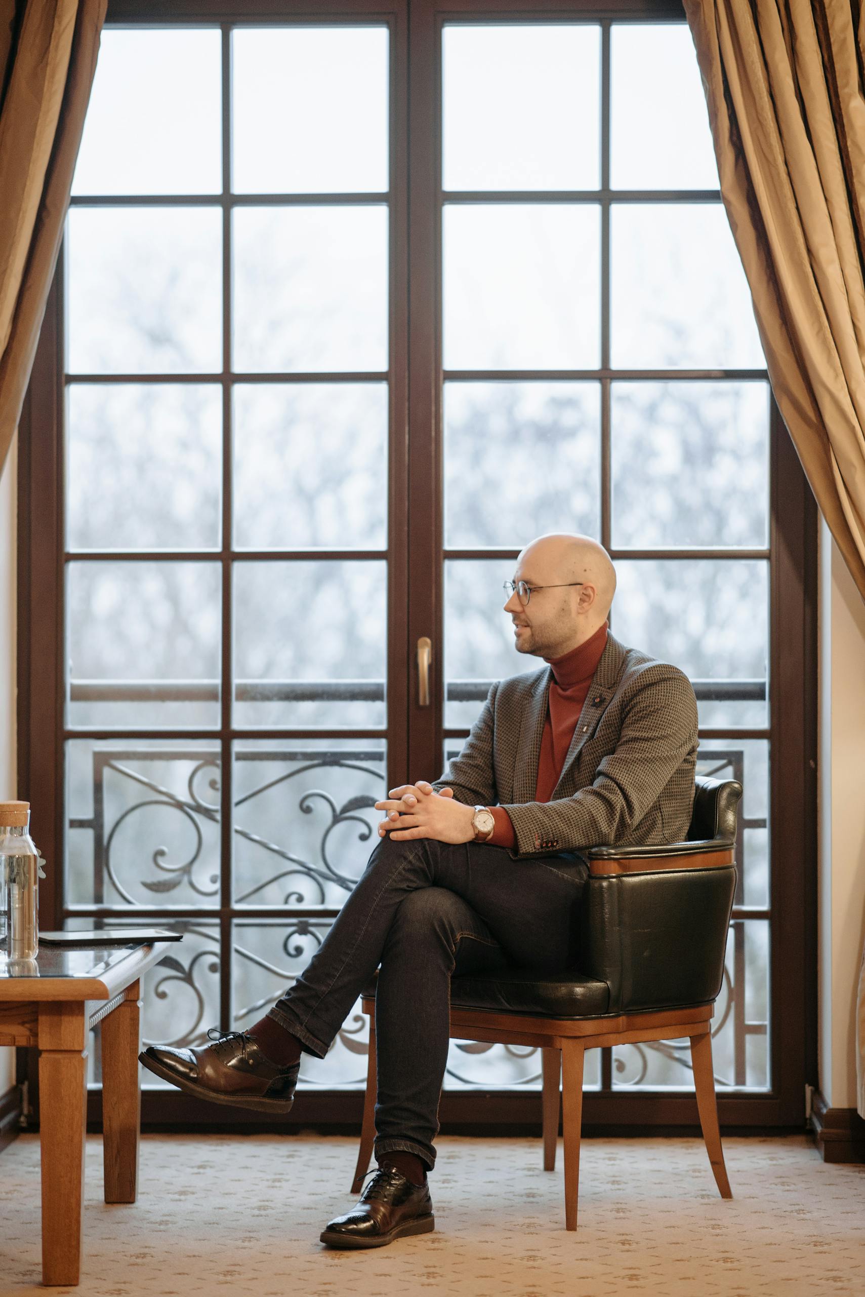 Man in a suit seated in an armchair by a large window indoors.